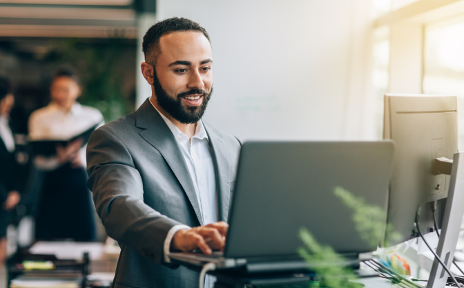 Foto de un hombre joven con barba sonriendo. vestido de traje gris  y leyendo en su computador portátil la información sobre protección de datos de fiduciaria Bancolombia.